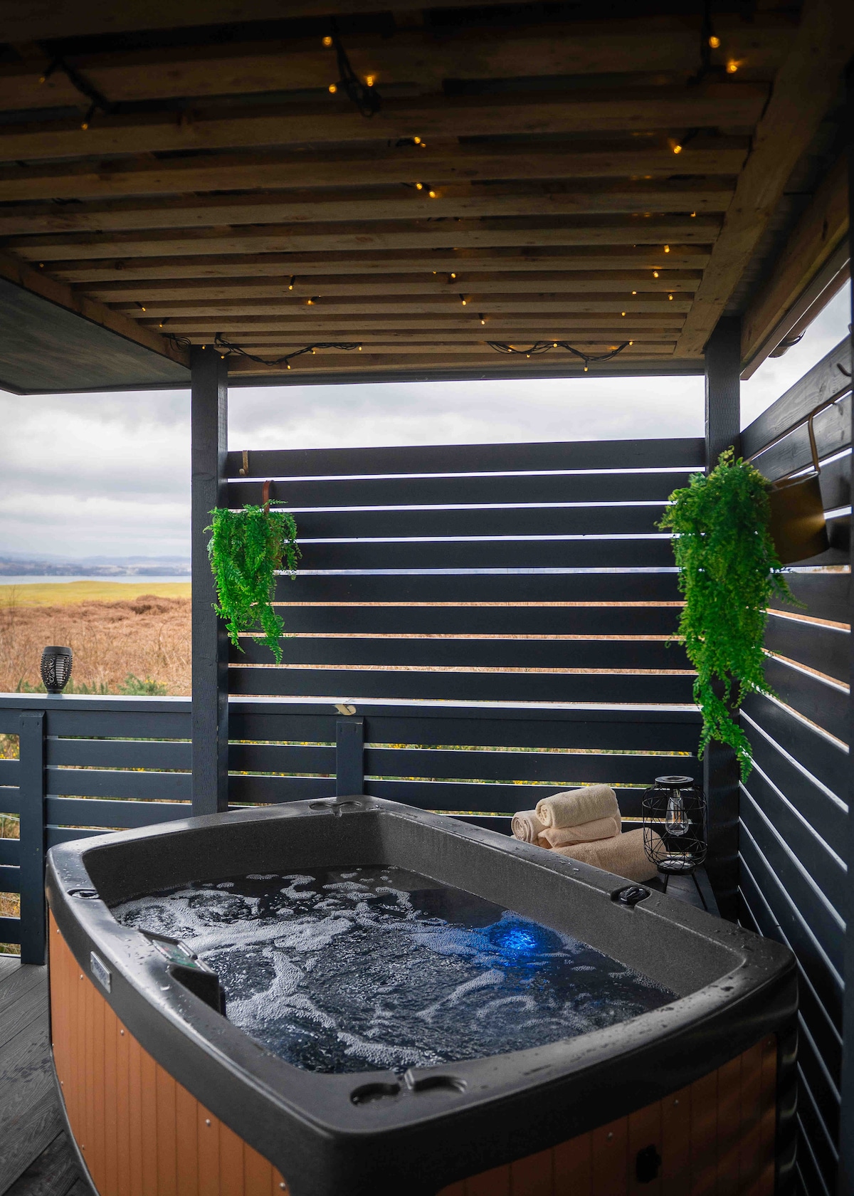 A hot tub is situated on a wooden deck, enclosed by dark wooden slats. Soft lighting illuminates the area, complemented by hanging green plants. Fresh towels are neatly arranged nearby, enhancing the overall sense of relaxation in this outdoor space.