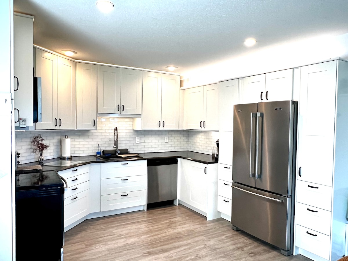 The kitchen features modern cabinetry with light tones and a clean design. Stainless steel appliances, including a refrigerator and dishwasher, are complemented by a black countertop. A white backsplash adds a bright touch, and the space is well-lit with recessed lighting.