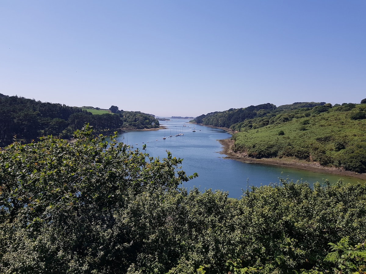 A serene view captures the winding Aber Benoît river, flanked by lush green hills and trees. Small boats can be seen on the gentle water, reflecting the clear blue sky. The image showcases the natural beauty of the surrounding landscape.