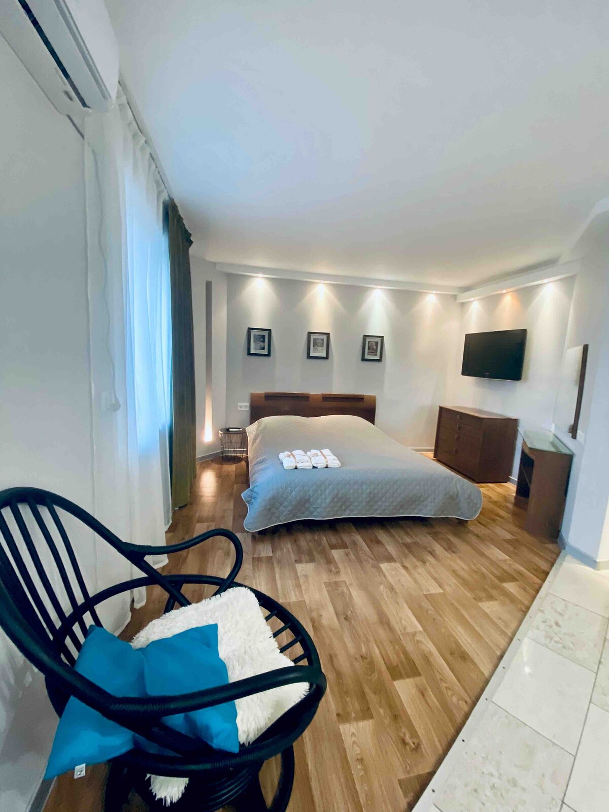 A spacious room features a queen-sized bed with a light gray quilt and decorative pillows. A black wooden chair with a blue accent pillow is positioned nearby. Ambient lighting highlights the walls, while a flat-screen TV and dark wood furniture enhance the contemporary decor.
