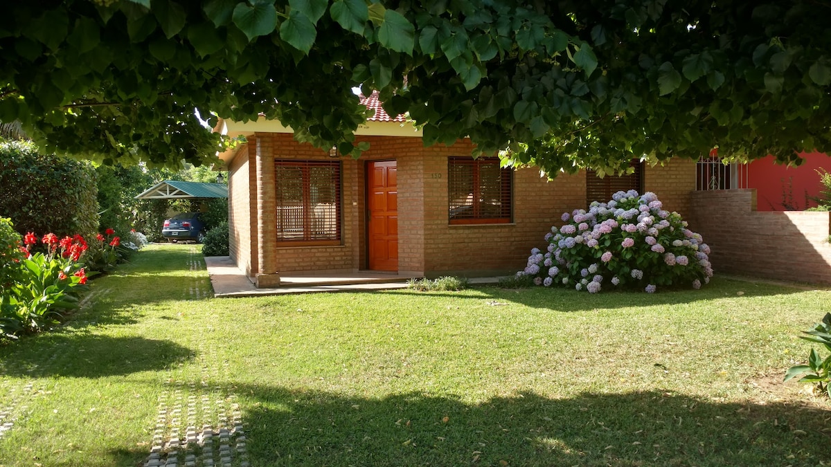 A single-story brick house is surrounded by a lush garden, featuring vibrant pink and red flowers. The entrance is framed by green foliage, and large windows offer ample natural light. A pathway leads to the house, contributing to the serene outdoor ambiance.