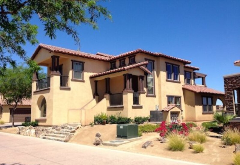 A split-level condo showcases a warm tan exterior with terracotta roof tiles. Large windows enhance the façade, allowing natural light to fill the spaces. Lush landscaping surrounds the property, featuring green plants and colorful flowers, contributing to the inviting atmosphere of the community.