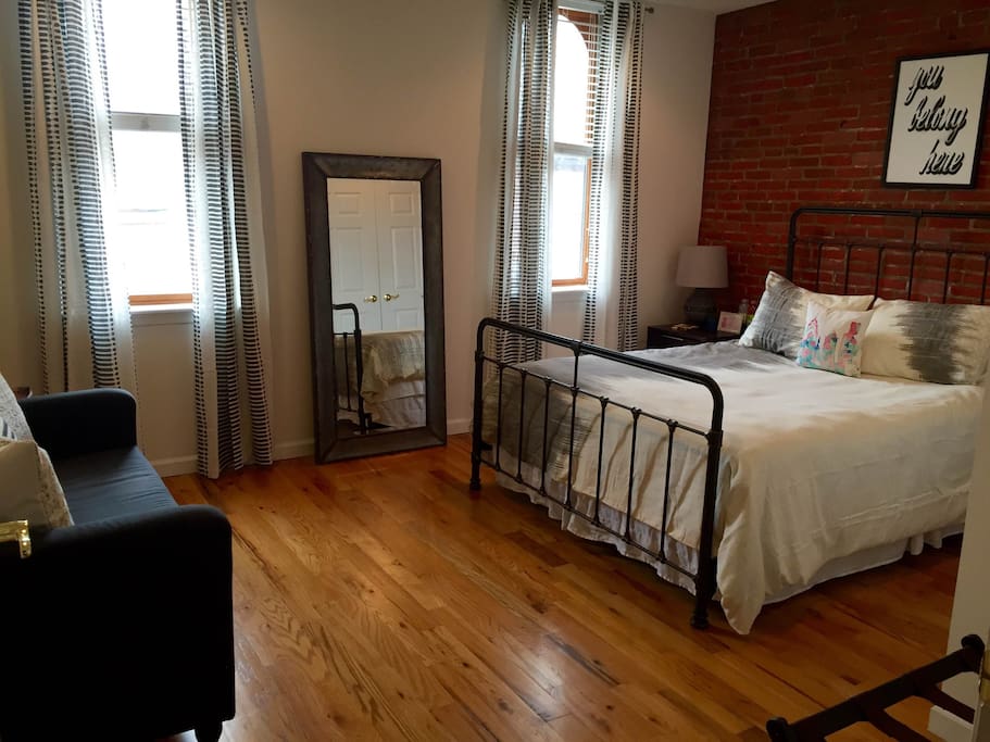 Welcome to your room: queen sized bed, exposed brick, a ceiling fan, and 2 large street-facing windows.
