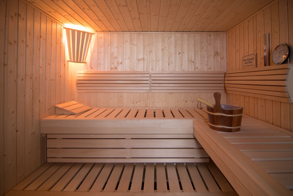 The sauna features a warm wood interior with an illuminated ceiling, providing a tranquil space for relaxation. Wooden benches are arranged along the walls, complementing a wooden bucket and ladle on the side. A thermometer and timer are visible, enhancing the wellness experience.