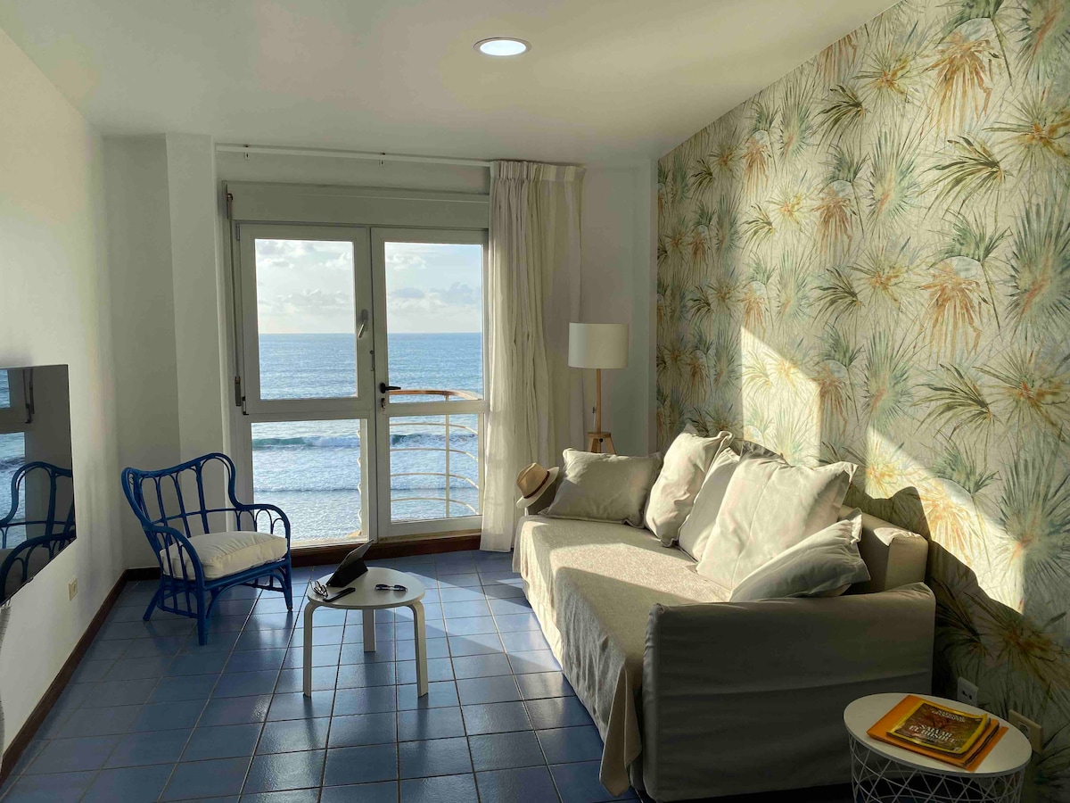 A bright sitting area features a comfy sofa with soft cushions, paired with a small white coffee table. A blue accent chair complements the space. Natural light floods in through large windows showcasing unobstructed ocean views, with a decorative wallpaper adding a touch of warmth.