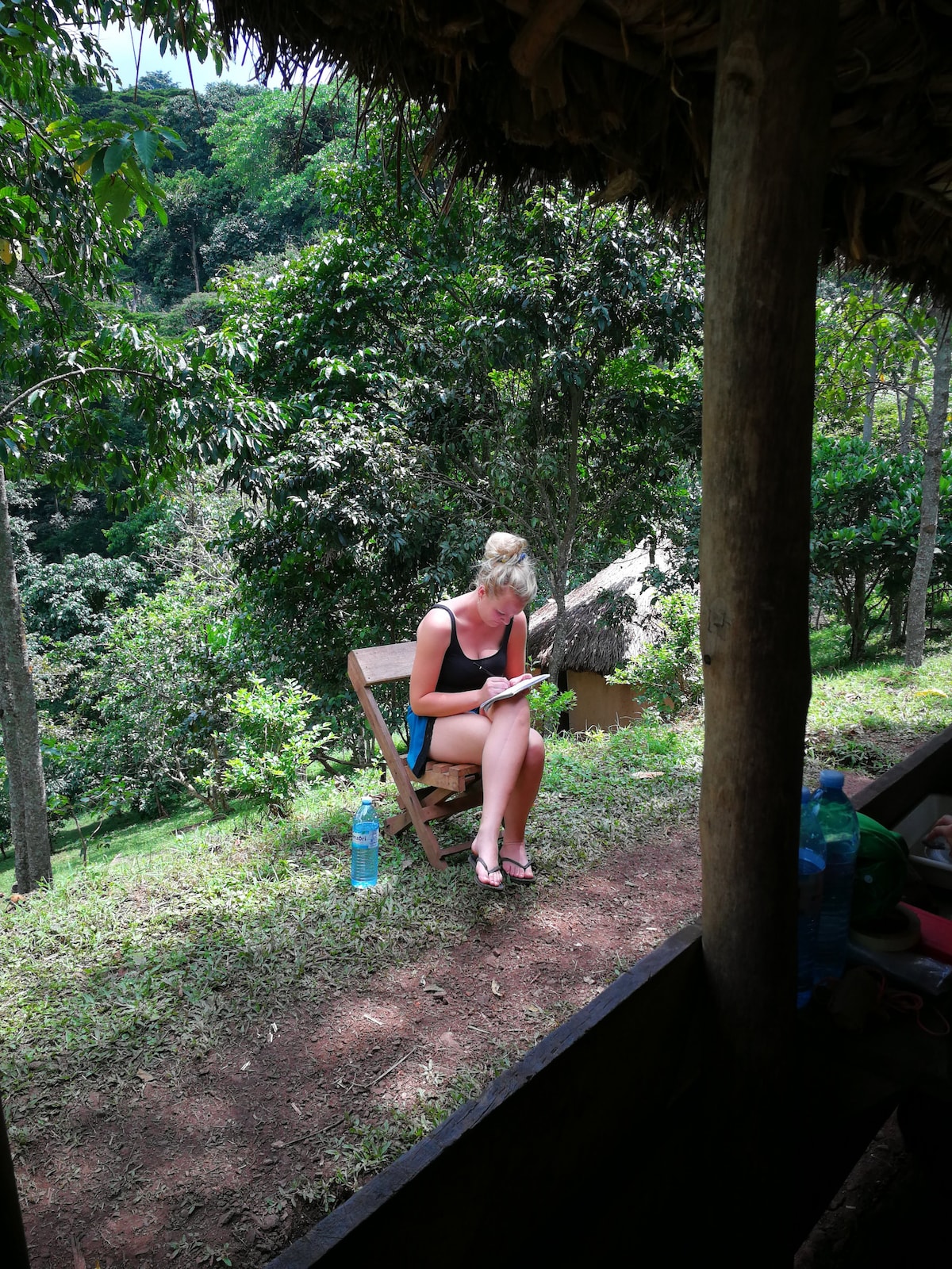 A wooden chair is positioned on a grassy area, surrounded by lush greenery. A person can be seen sitting and enjoying a snack, with a bottle of water beside them. In the background, natural vegetation and a small structure are visible through the trees.