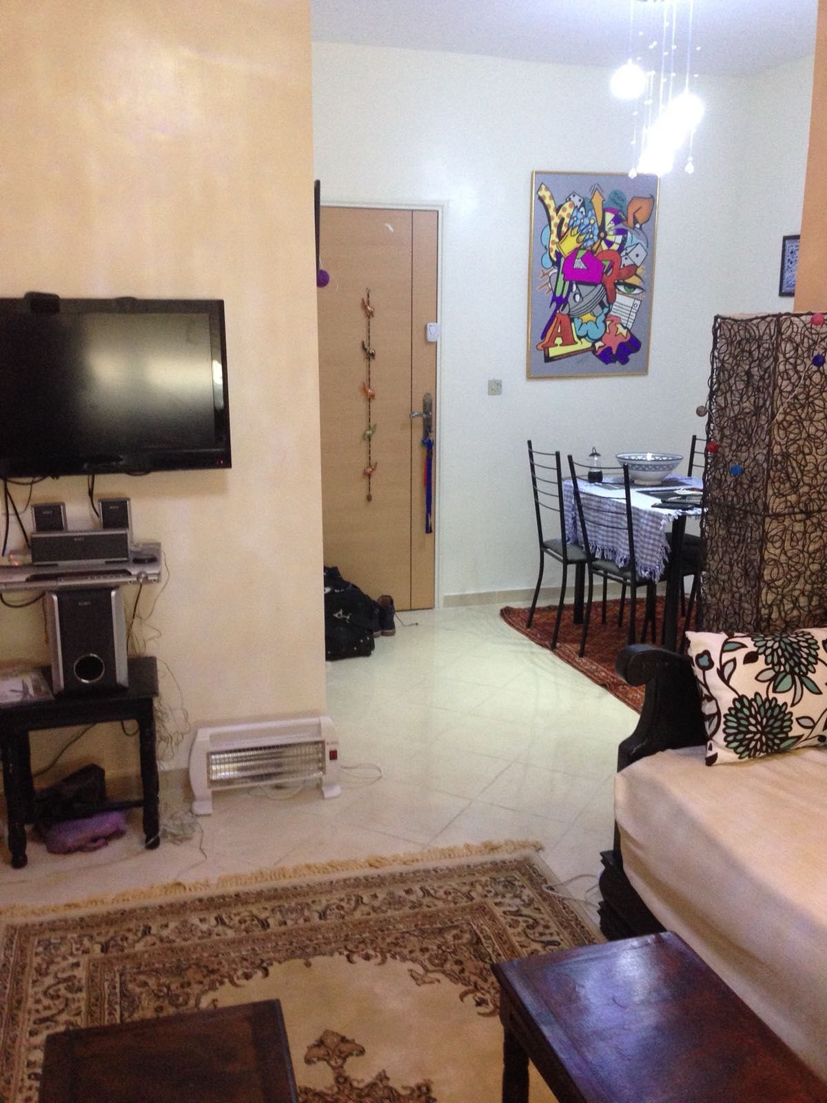 An inviting living area is observed, featuring a comfortable sofa and decorative elements. A television is mounted on the wall, while a dining space with a table and chairs is visible in the background. Colorful artwork hangs on the wall, adding character to the room.