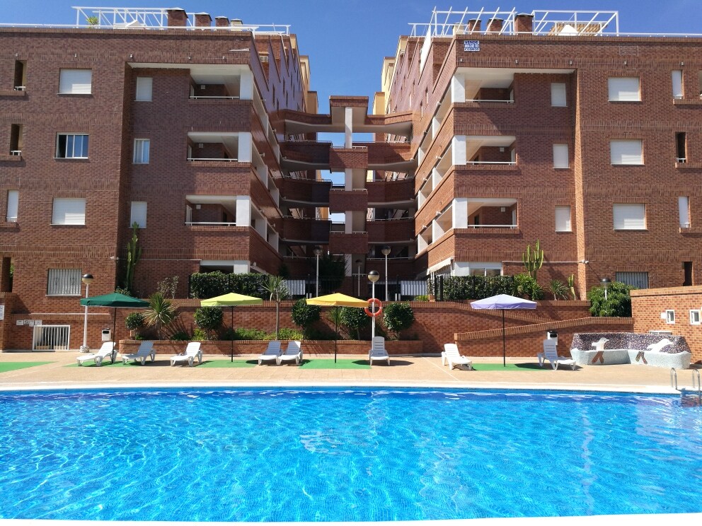 The photo shows a multi-story brick building adjacent to a vibrant pool area. Sun loungers are arranged by the water, while umbrellas in various colors offer shade. Lush greenery and landscaped gardens complement the serene environment, creating a welcoming atmosphere for relaxation.