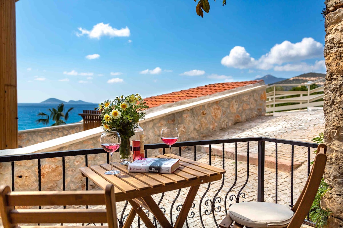 A private terrace offers a small wooden table set with two glasses and a bottle of wine. Vibrant flowers are showcased in a vase, with the sea visible in the background under a bright blue sky. Stone walls and a tiled roof frame the scene.