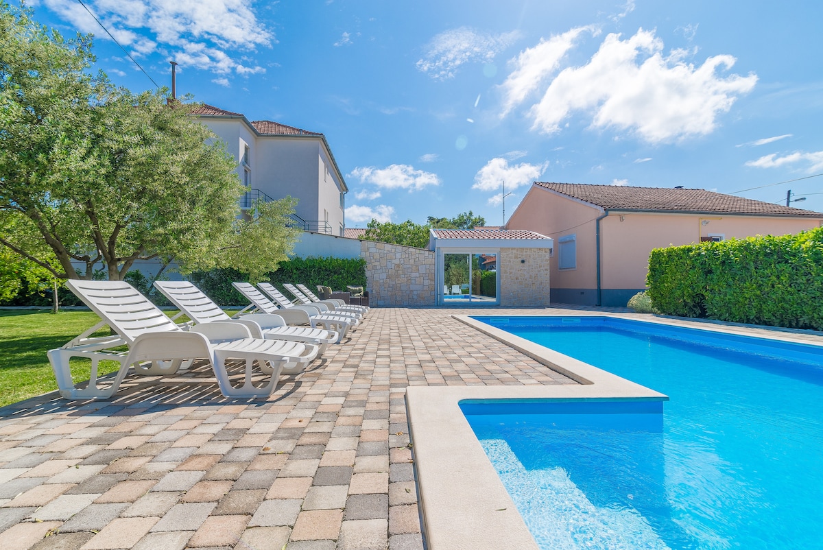 An outdoor pool surrounded by a sun terrace features multiple lounge chairs arranged neatly along the edge. Lush greenery and trees provide shade, while a building in the background complements the tranquil atmosphere. The clear blue water reflects a bright sky dotted with clouds.