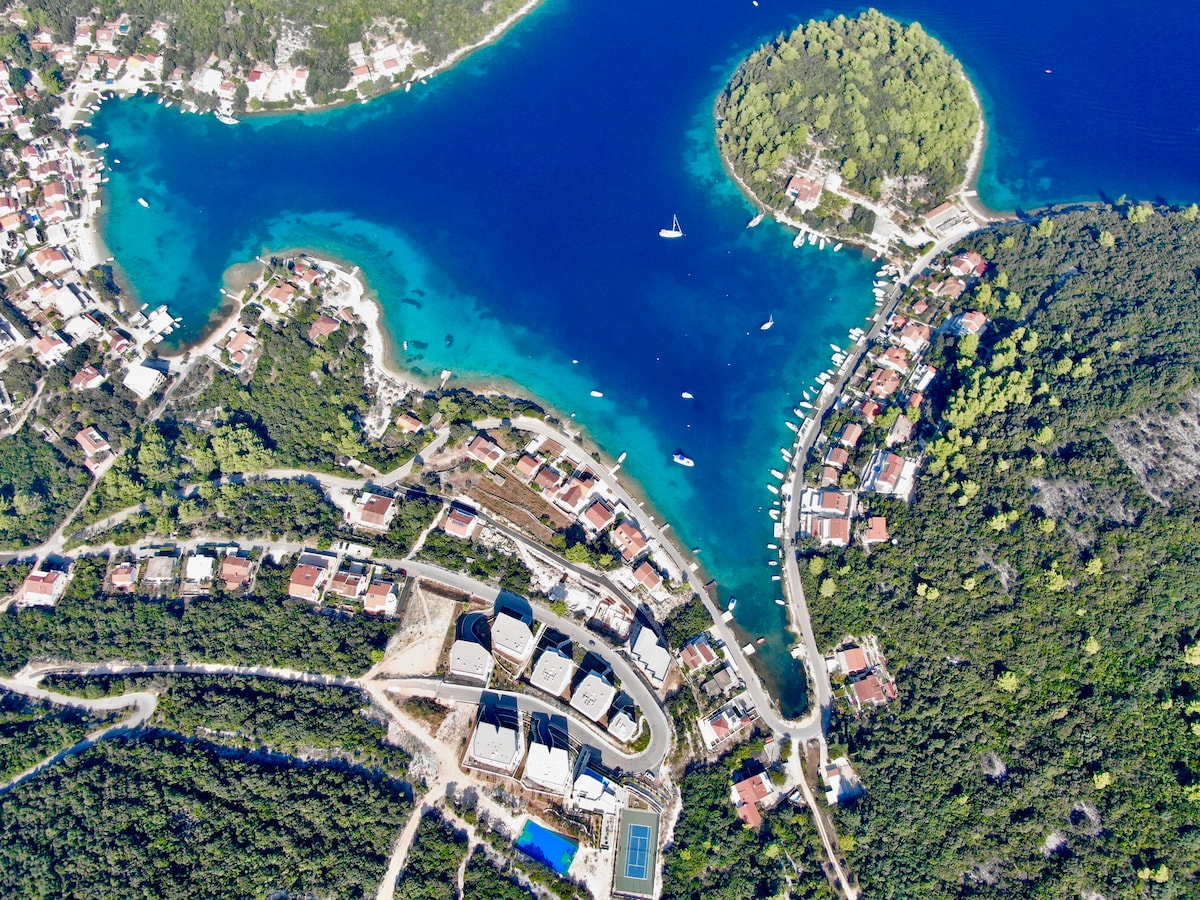 An aerial view showcases the vibrant blue waters of Žrnovska Banja, surrounded by green coastal hills and residential buildings. The layout features a mix of boat docks and winding roads leading to the luxurious Blue Bay apartments, highlighting the natural beauty and tranquility of the area.