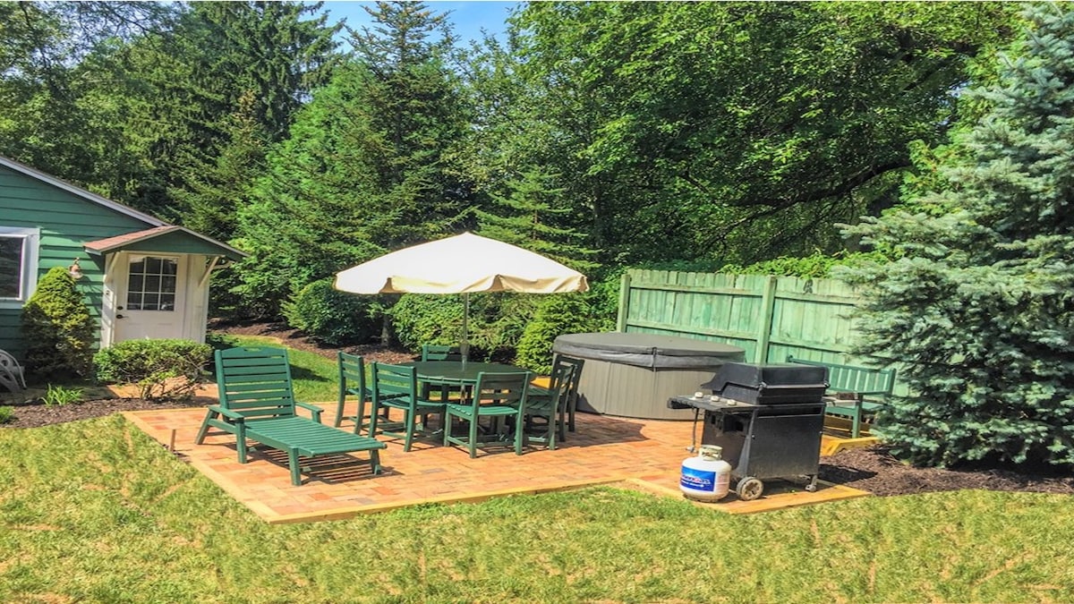 An outdoor patio area is depicted featuring green lounge chairs and a dining set arranged on brick pavers. A large umbrella offers shade over the seating. A hot tub is positioned nearby, surrounded by tall greenery, complemented by a grill against a fenced backdrop.