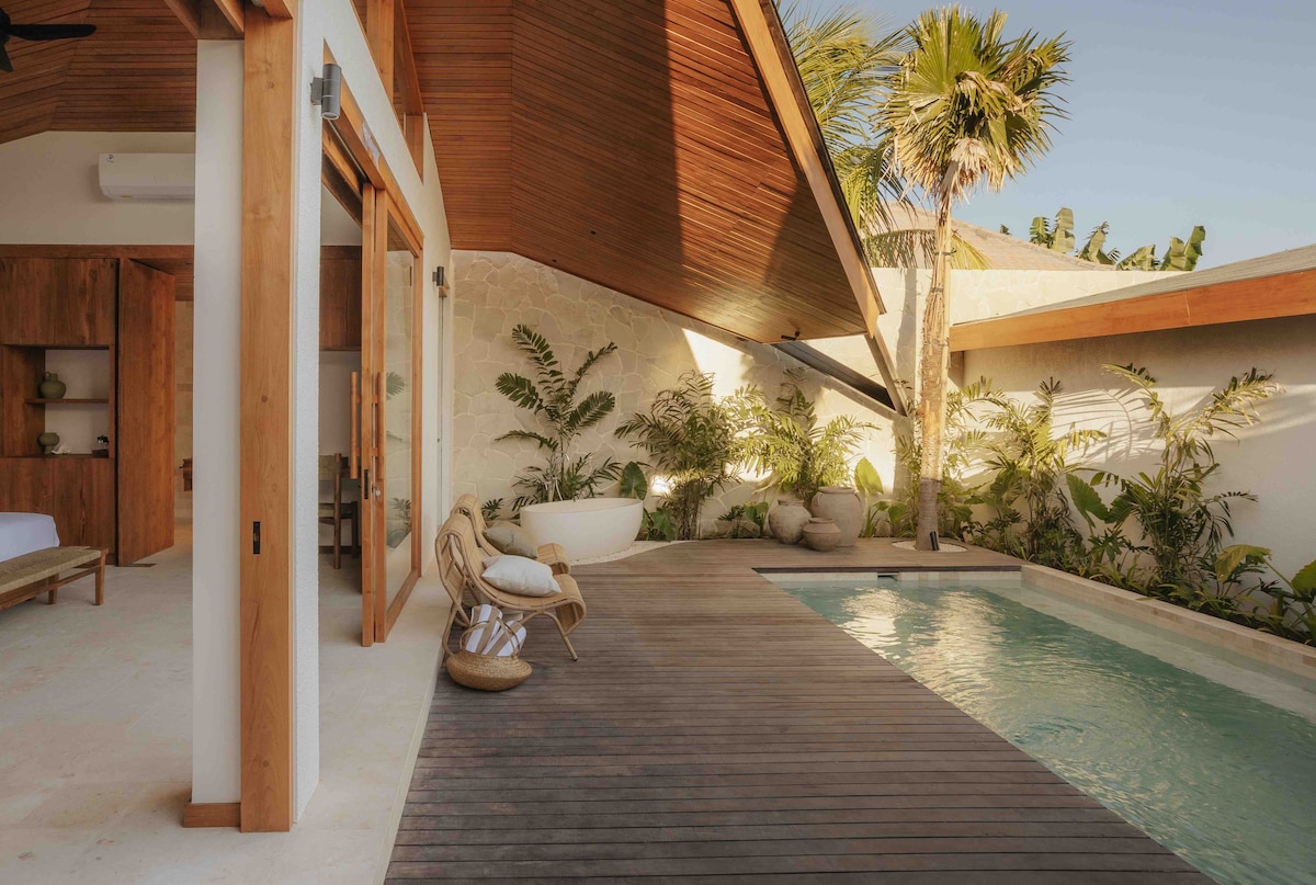 A private pool is surrounded by a wooden deck and tropical plants, creating a serene outdoor space. The architectural design features high ceilings with wooden beams. An outdoor bathtub is positioned near the edge of the pool, complementing the tranquil setting.