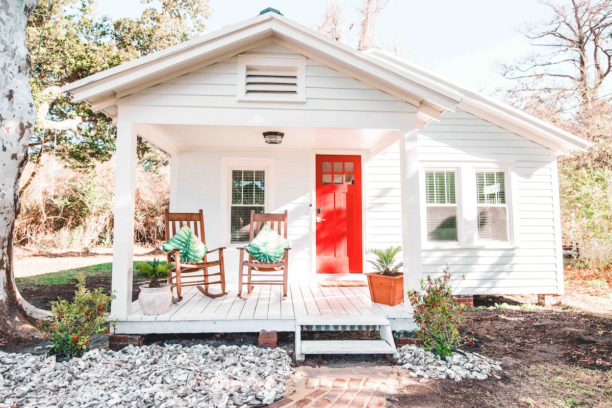 A charming bungalow exterior is shown, featuring a red front door and a welcoming porch with two rocking chairs adorned with green cushions. Surrounding greenery and well-maintained landscaping complement the home's inviting appearance.