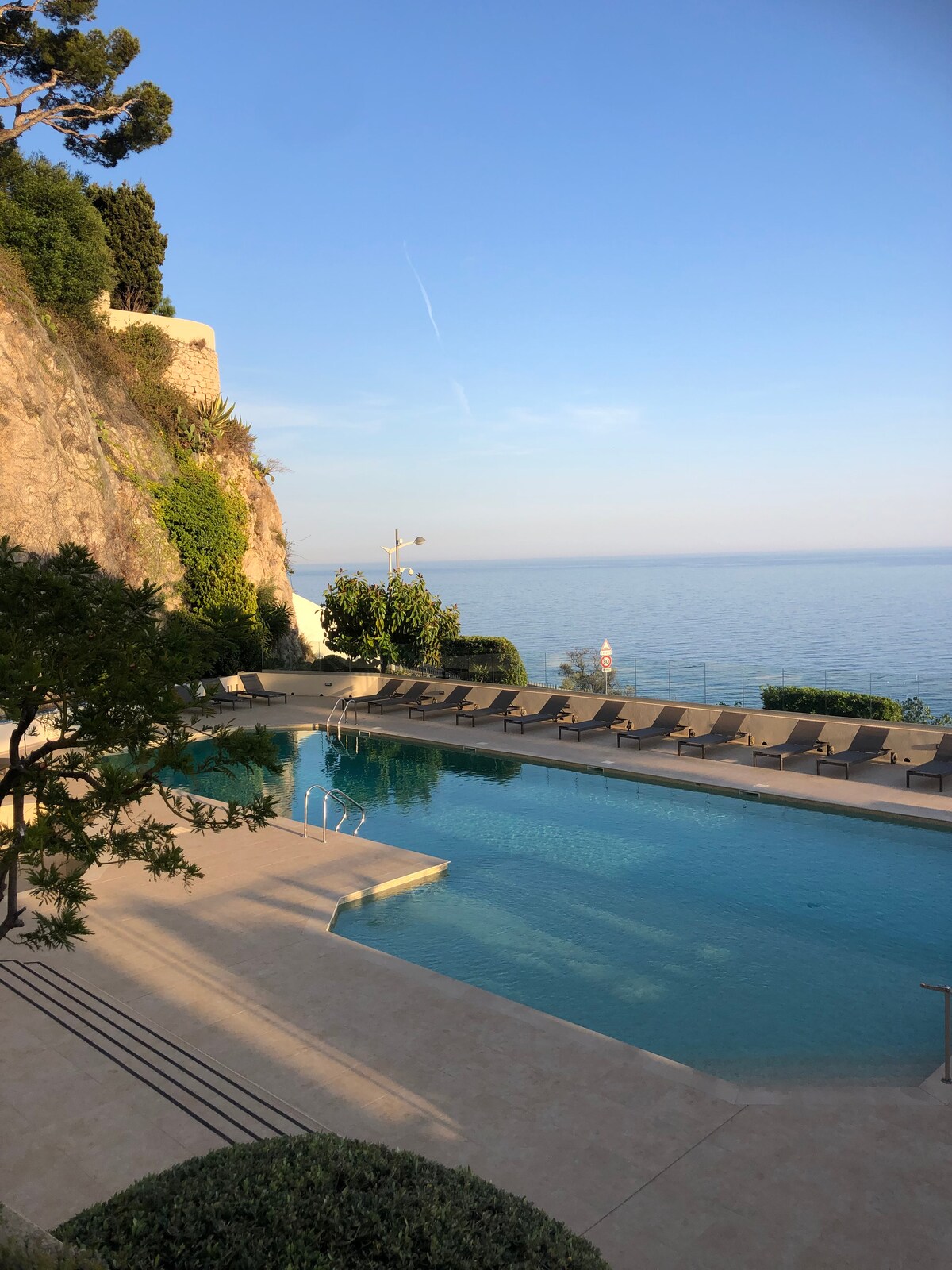A serene swimming pool is set against a backdrop of a rocky hillside, with lounge chairs lined along the water's edge. The pool reflects the calm sea, offering a tranquil atmosphere and unobstructed views of the horizon.