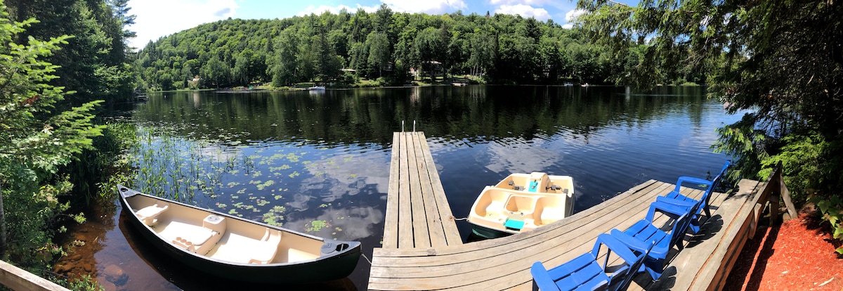 A serene lakeside view is presented, showcasing a calm water surface reflecting surrounding trees. A wooden dock extends into the lake, with two boats positioned alongside. Brightly colored chairs sit on the dock, inviting relaxation near the water's edge.