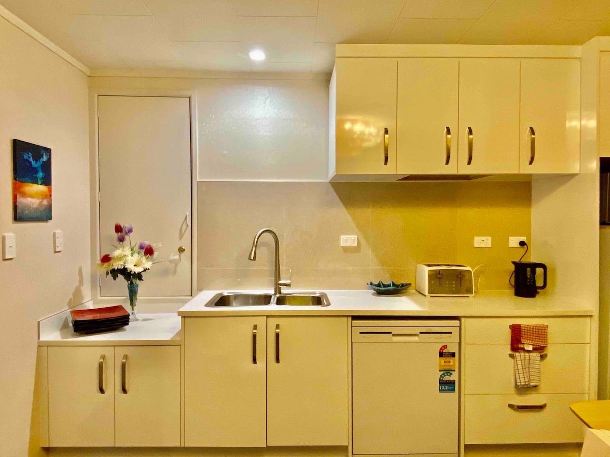 The modern kitchen is equipped with a stainless steel sink and sleek cabinetry. A microwave, kettle, and a compact refrigerator are visually accessible. Decorative items and a small window add a touch of color. The countertop offers ample space for meal preparation.