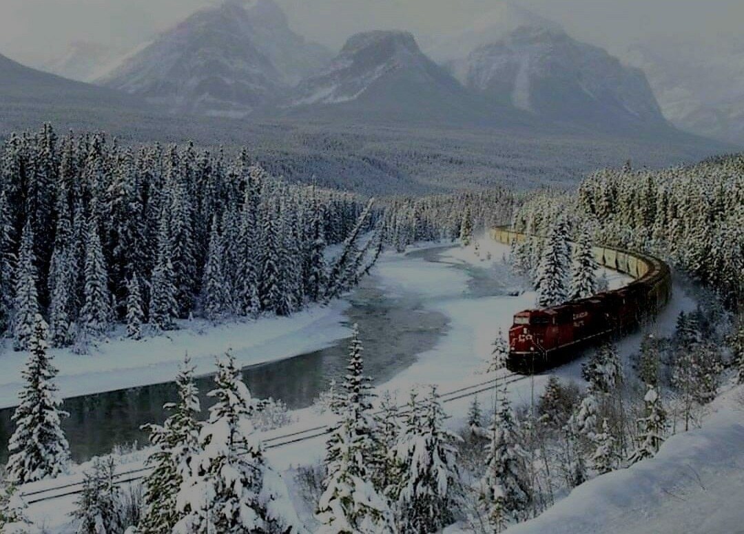 A train travels along a snow-covered landscape, next to a winding river surrounded by evergreen trees. Snow-capped mountains rise in the background, creating a serene and quiet winter scene.