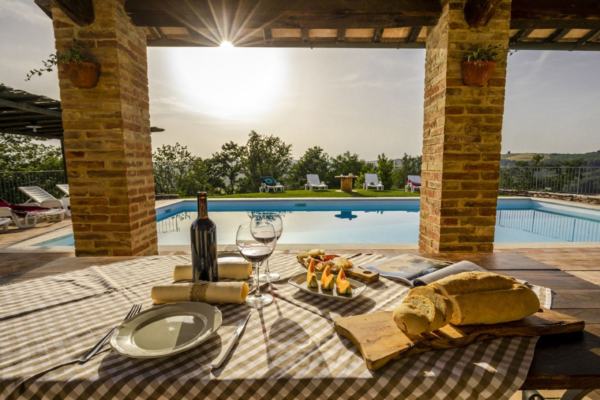 A dining table is set with plates and wine glasses, accompanied by bread and appetizers. The scene is framed by stone columns, with sunshine casting a glow over the inviting swimming pool and surrounding loungers in the background.