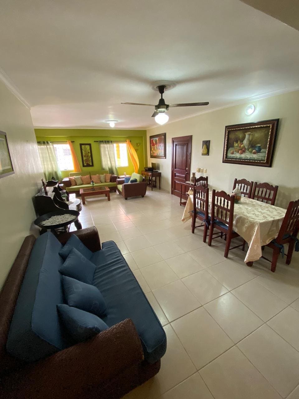 The living area features a mix of seating options, including a brown couch with blue cushions and green armchairs. A dining table with wooden chairs accommodates several guests. Natural light enters through large windows, partially covered by sheer drapes, complementing the green walls and decorative artwork.