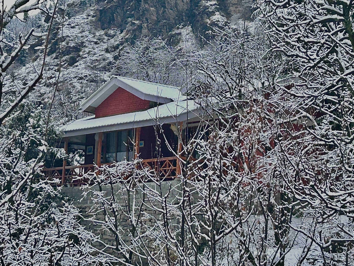 The independent cottage is nestled among snow-covered trees, featuring a red brick exterior and an extended porch. A backdrop of rocky terrain and frosted foliage highlights the serene atmosphere of this winter landscape.