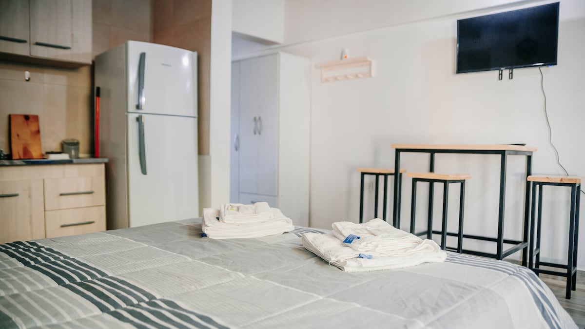 A neatly arranged sleeping area features a spacious bed covered with a light grey blanket. Fresh towels are placed at the foot of the bed. In the background, a kitchen area with a refrigerator and dining stools is visible, alongside a mounted television.