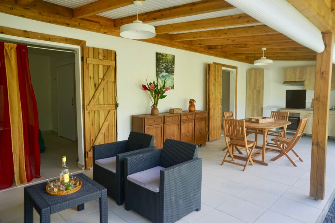 A spacious living area features natural wood beams and large outdoor access. Comfortable seating with dark wicker chairs is arranged around a small table. A wooden dining table with folding chairs is positioned nearby, creating an inviting space for relaxation and meals.