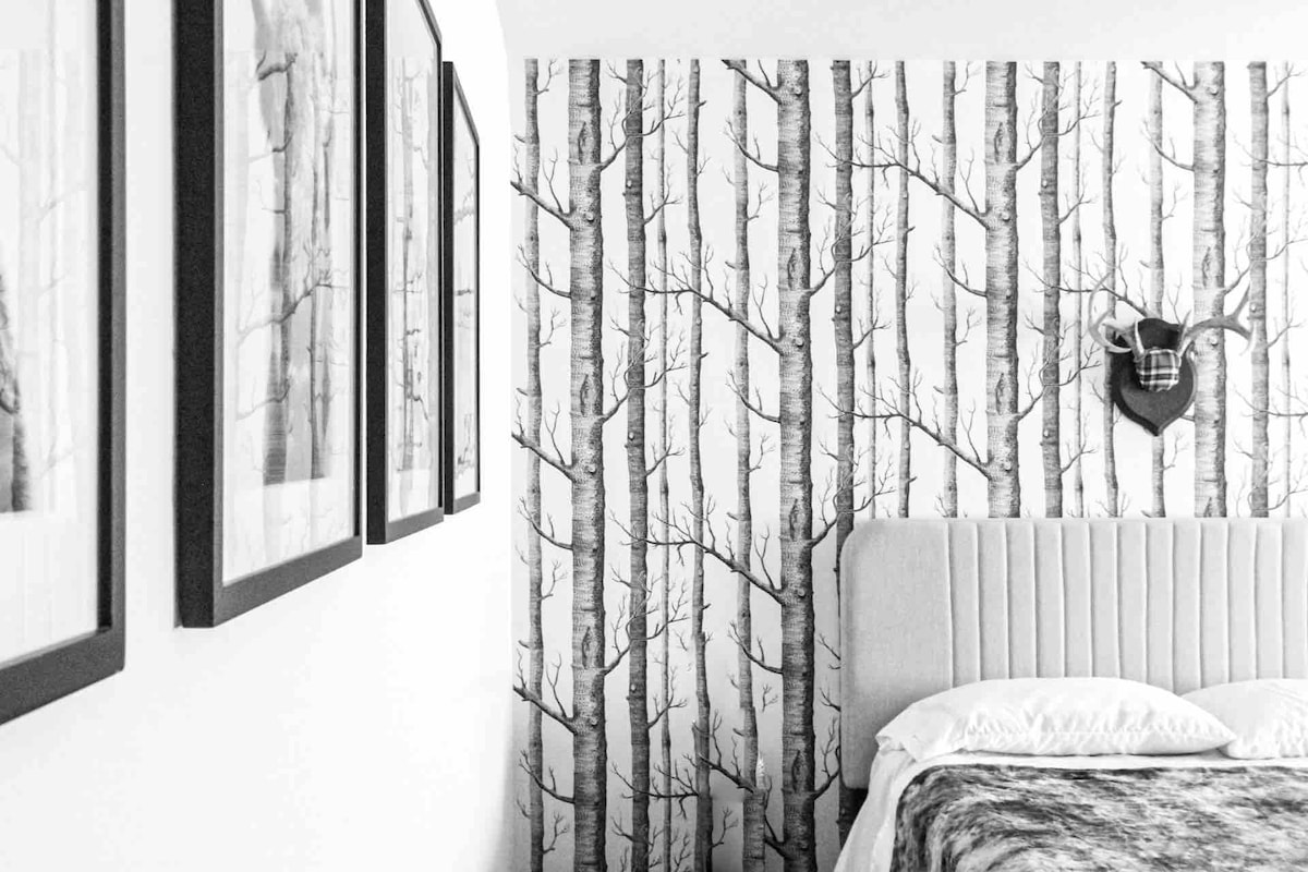 A modern mountain-themed bedroom features a soft-toned headboard positioned against a birch tree wallpaper. Framed black-and-white photographs hang on the wall, and a decorative antler piece adds a rustic touch above the bed, enhancing the serene ambiance.