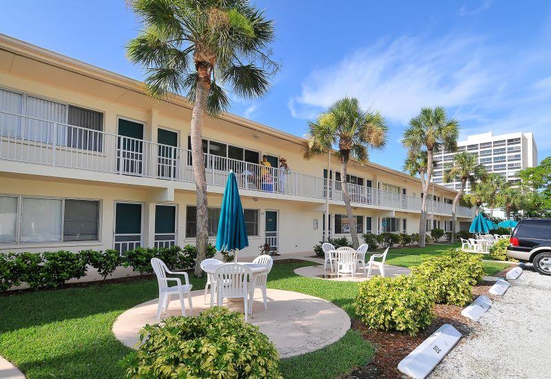 The exterior of the two-story condo building is visible, featuring a series of balconies adorned with umbrellas. Lush greenery surrounds circular seating areas with white chairs and tables, providing a relaxed outdoor atmosphere. Clear blue skies complement the setting.
