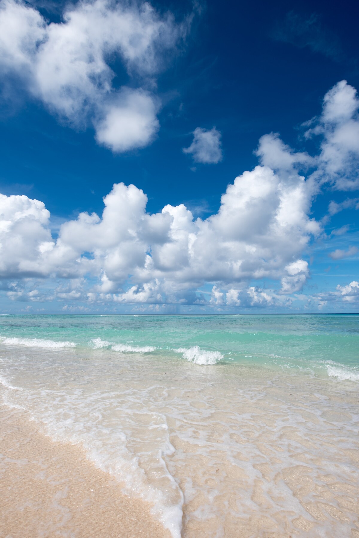 A serene beach scene features soft, golden sand meeting calm, turquoise waters. Dynamic clouds float above against a clear blue sky, creating a tranquil atmosphere. Gentle waves lap at the shore, inviting relaxation and enjoyment of the natural beauty.