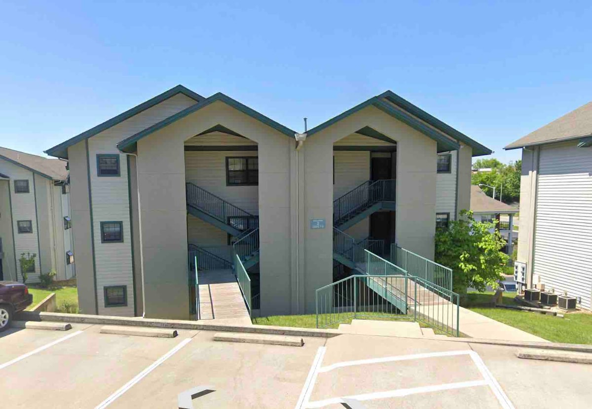 The exterior of a two-story condominium complex is captured, showcasing a light-colored facade with green accents. A staircase can be seen leading to the second level, bordered by railings. Parking spaces are visible in the foreground, with greenery framing the scene.
