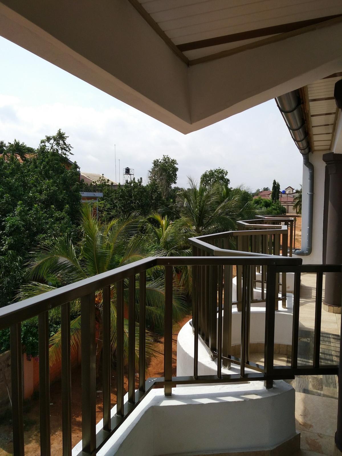 A spacious terrace features a brown railing and curves around a corner, overlooking lush greenery and palm trees. Natural light brightens the area, creating a serene atmosphere that invites relaxation and contemplation.