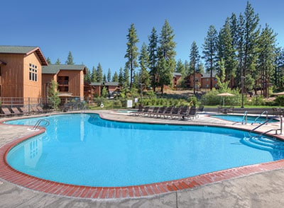 A spacious outdoor pool is surrounded by trees and buildings, featuring a clear blue water surface. Several lounge chairs are arranged along the poolside, inviting relaxation under the sun. The deck is finished with a textured stone surface, enhancing the inviting leisure space.