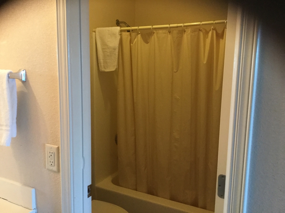 A clean bathroom area is showcased, featuring a shower/tub combination with a neutral shower curtain. Soft white towels are hanging on the nearby towel rack, and the sink area is presented with a simple counter space. The light beige walls create a calming environment.