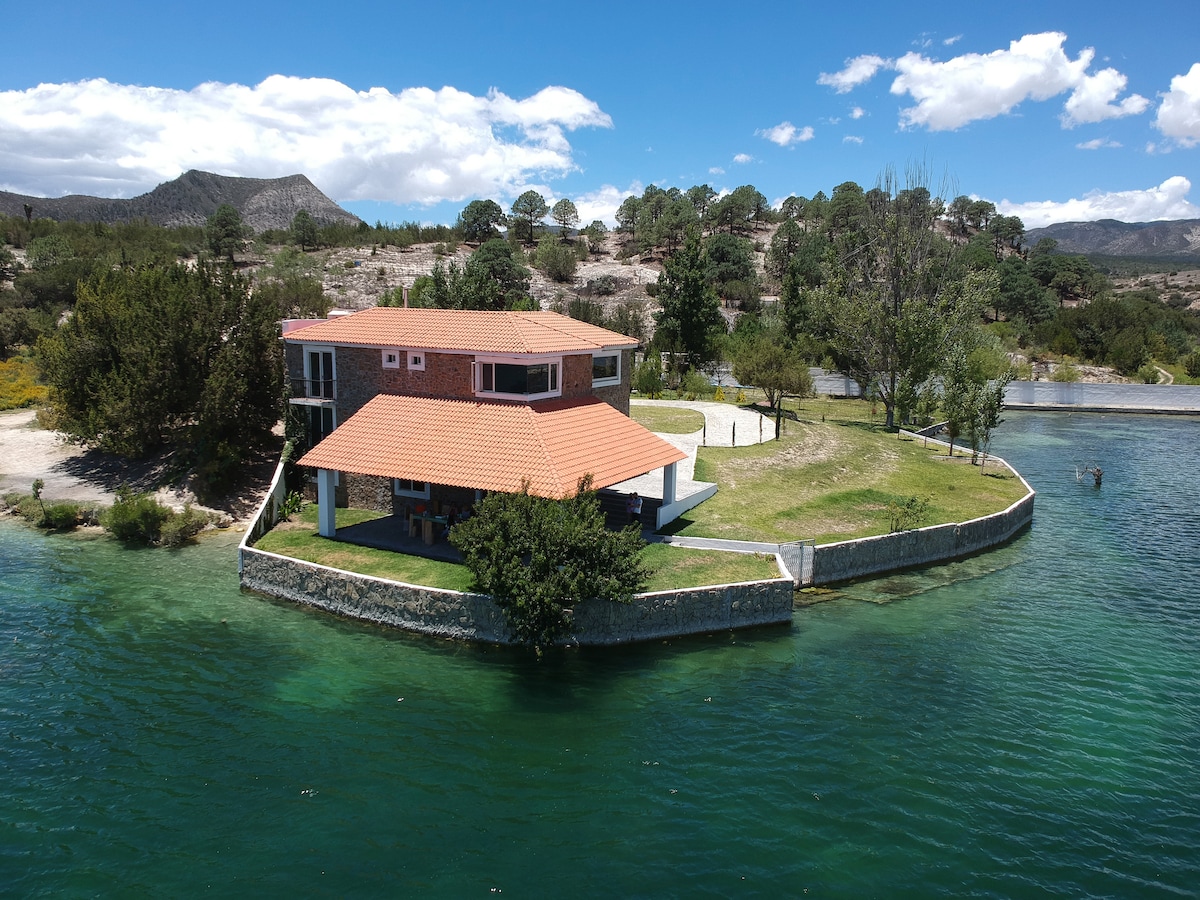 A spacious house is positioned on a peninsula surrounded by clear turquoise waters. The building features a red-tiled roof and green outdoor spaces, with a backdrop of scenic hills and pine trees. The site is set in a tranquil, natural environment.