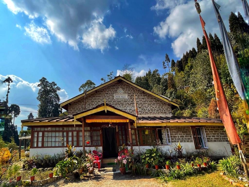 The heritage bungalow is framed by lush greenery, showcasing its solid stone masonry exterior. The spacious front porch is adorned with potted plants and chairs, inviting relaxation. Large windows allow natural light to permeate the interior, while mountains can be seen in the background.