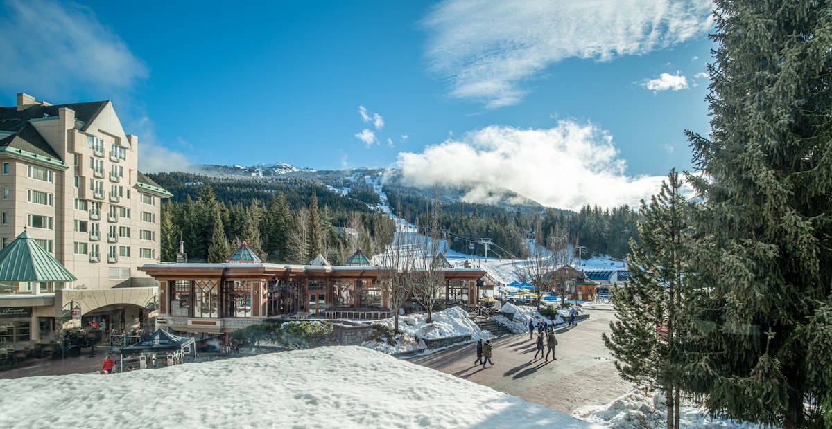 An outdoor winter scene captures the bustling Upper Village, accompanied by snow-covered grounds. The backdrop features skiers on nearby slopes and trees surrounded by a clear blue sky. The village area is lively with people walking, and various buildings are visible, including a hotel with a distinct green roof.