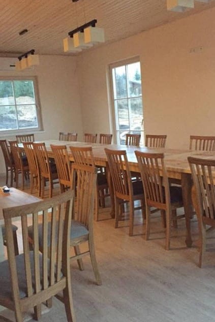 A spacious dining area features a long wooden table surrounded by multiple wooden chairs. Large windows allow natural light to fill the room, while the simple, neutral walls create an open atmosphere. Modern light fixtures hang from the ceiling, adding a touch of sophistication.