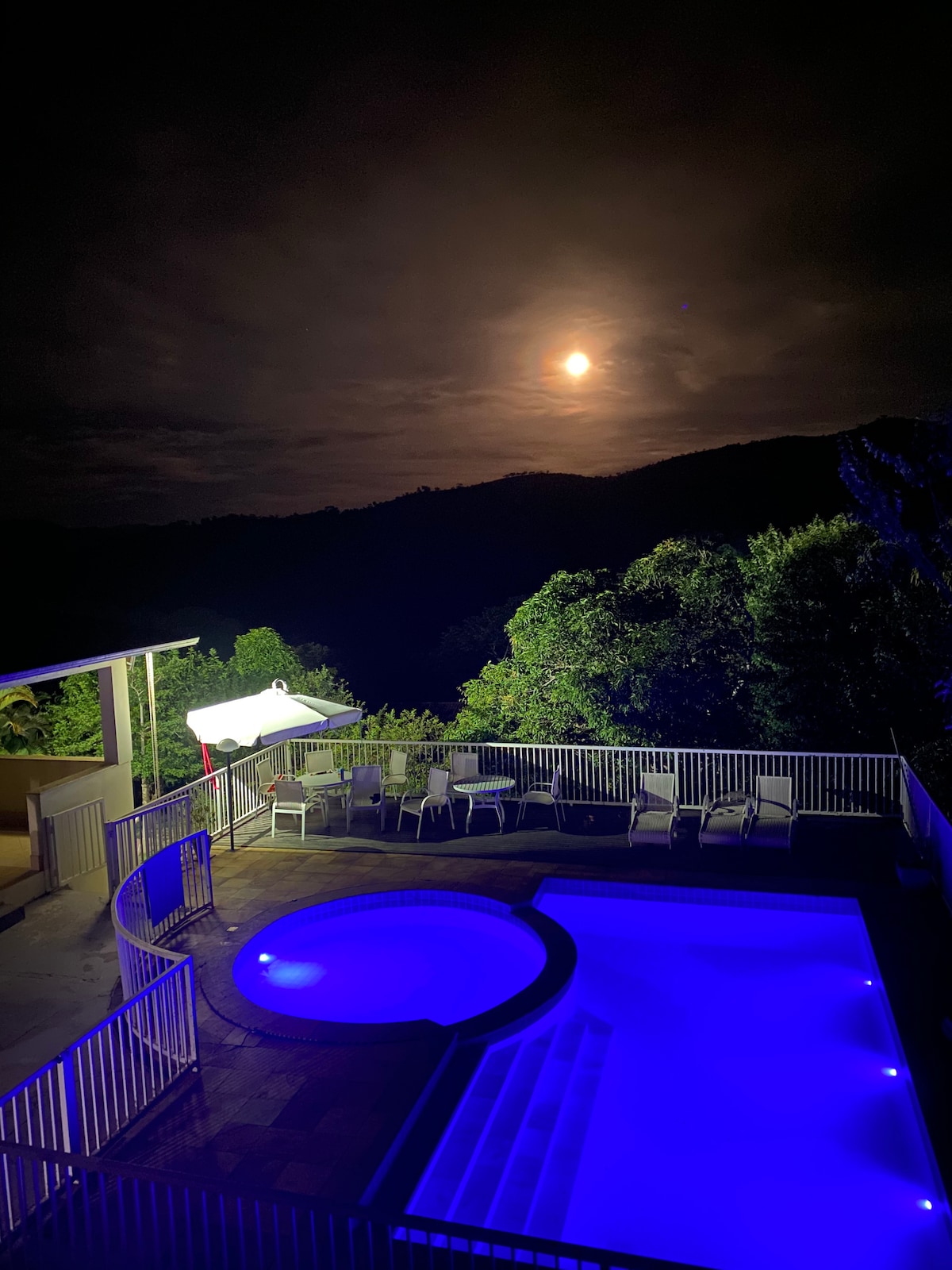 The scene captures a serene night view of a lit pool area with a circular jacuzzi. Surrounding greenery complements the ambiance, while a full moon casts a soft light over the setting. A white umbrella and several chairs are visible around the pools.