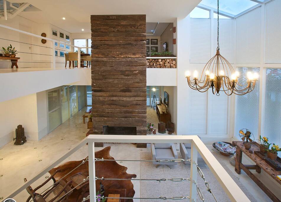 O living room da residência se destaca pelo espaço amplo de pé-direito duplo e com presença abundante da luz natural que invade o interior da casa através das amplas aberturas de vidro. O mobiliário de marcenaria, o lustre de estilo retrô e o amplo painel revestido de dormentes dão um toque rústico ao ambiente e realçam a sensação de aconchego.