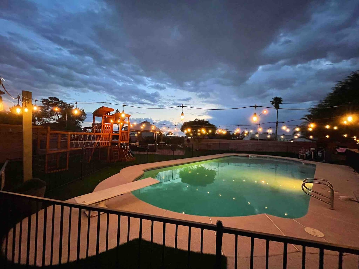 A spacious backyard is highlighted by a large pool, surrounded by a fenced area. Soft lights are seen strung above, creating a warm ambiance. A children's playset is positioned nearby, complimenting the relaxing atmosphere enjoyed during the evening.