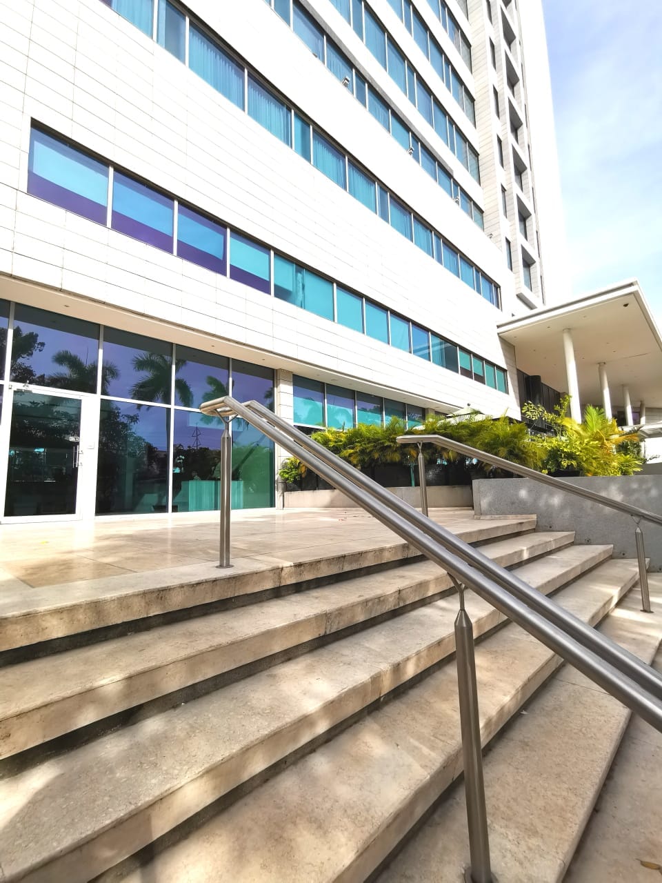 A modern building entrance is framed by a series of wide stone steps and gleaming glass doors. The exterior features sleek lines and large windows, allowing natural light to flood the interior. Lush green plants are arranged along the base for added greenery.