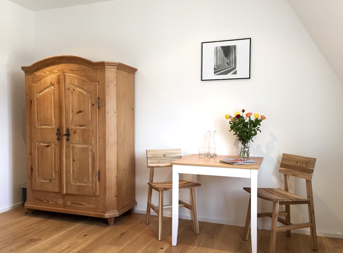 A wooden armoire stands against a white wall, showcasing a simple design. A small table with two matching chairs is set nearby. A vase of fresh flowers adds a touch of color, complemented by a framed black-and-white photograph hanging above.