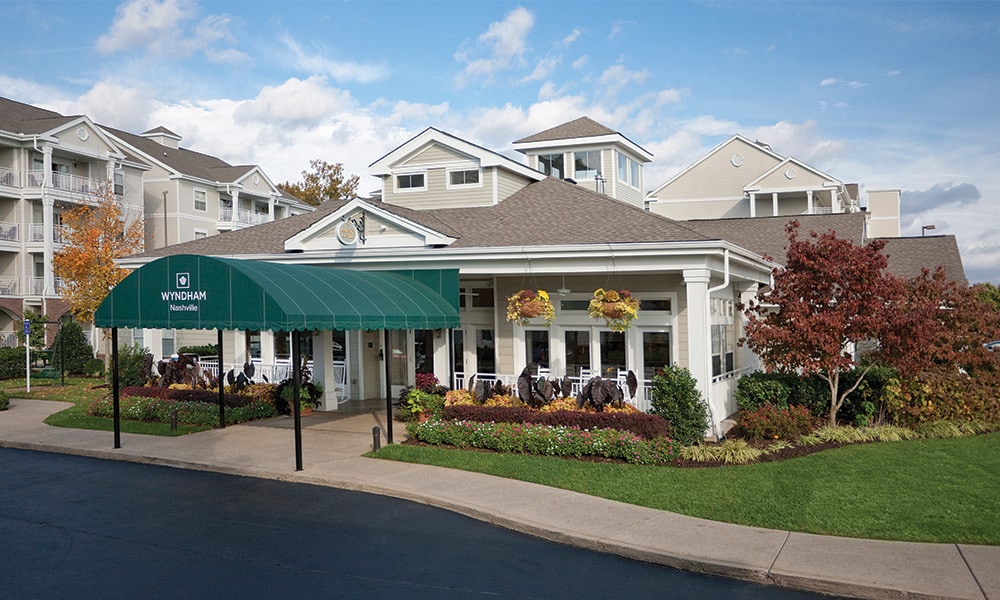 The resort entrance features a decorative green awning, welcoming guests. Surrounding the entrance is a landscaped area with seasonal flowers and shrubs. The two-story buildings with light-colored exteriors create a charming atmosphere, set against a partly cloudy sky.