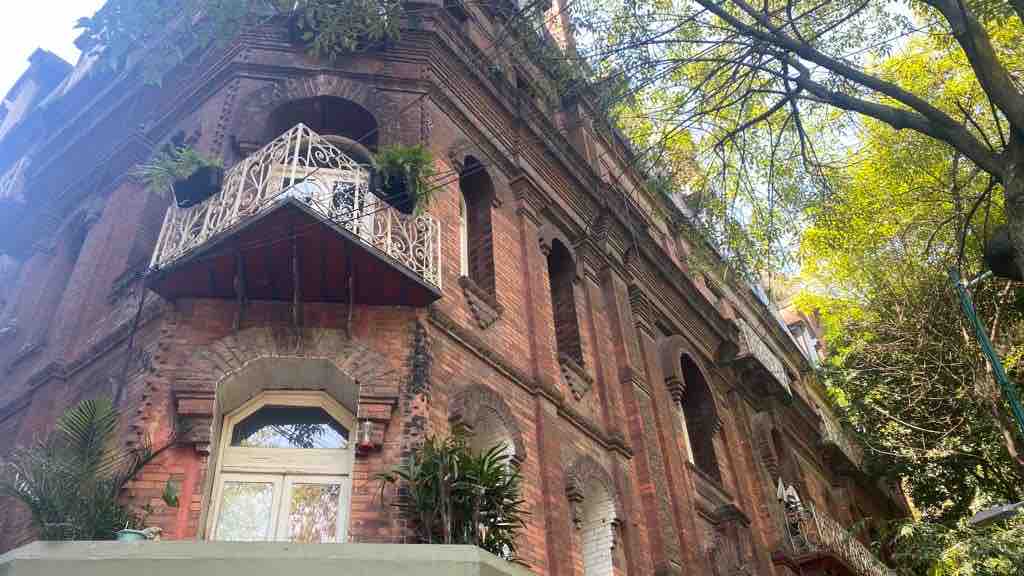 The building features intricate brickwork and ornate balconies adorned with greenery. Tall windows allow natural light to enter, while the surrounding lush trees create a serene environment. The architectural details reflect a historic charm in the neighborhood.