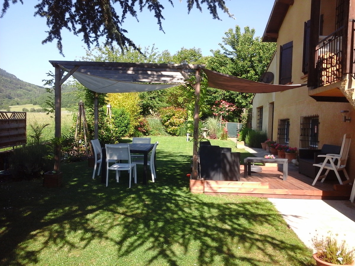 A private garden is shown, featuring a shaded patio with a wooden table and chairs. A lounge area with comfortable seating is visible, surrounded by well-maintained greenery. The landscape includes hills and trees in the background, creating a peaceful outdoor setting.
