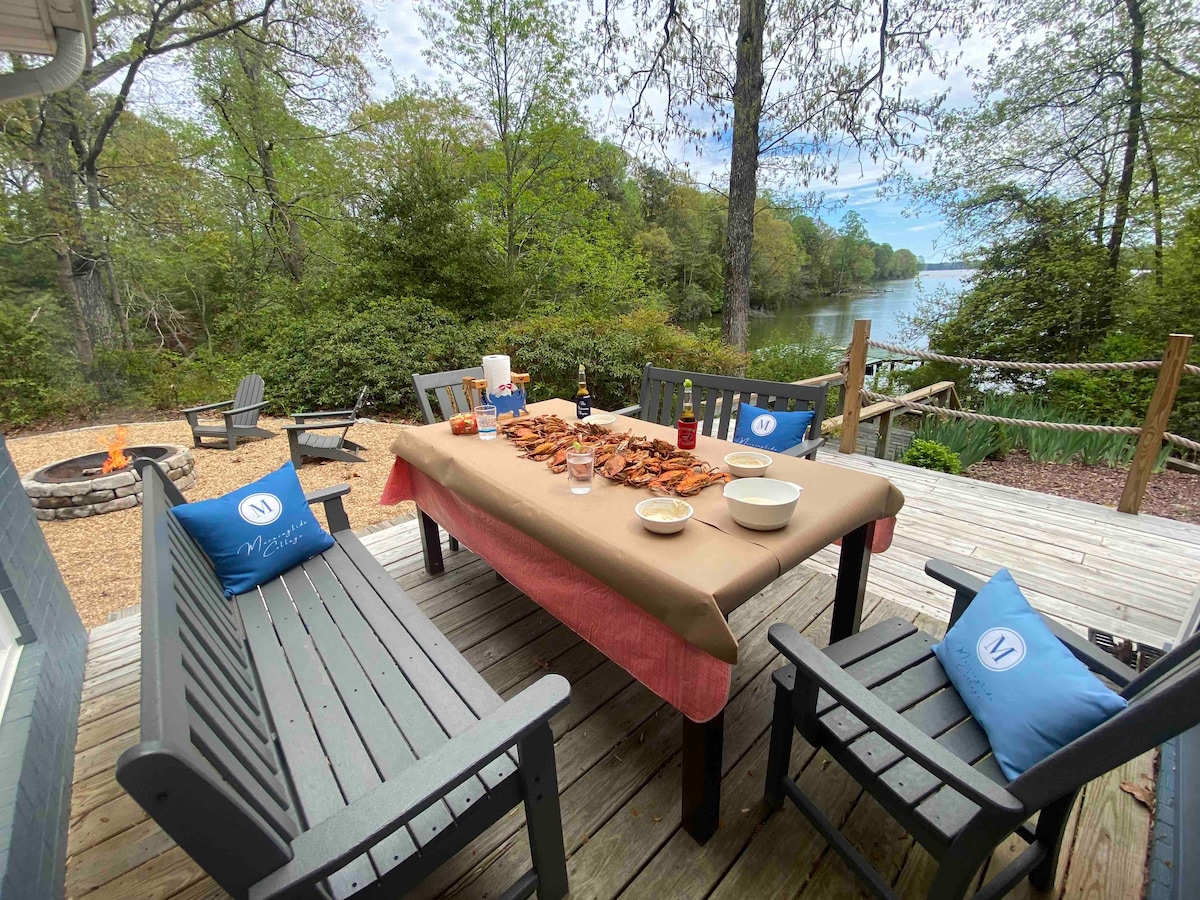 An outdoor dining area is set on a wooden deck, facing a serene waterfront view. A table is arranged with bowls and beverages, surrounded by comfortable seating. A fire pit is visible nearby, complemented by lush greenery and trees framing the scene.