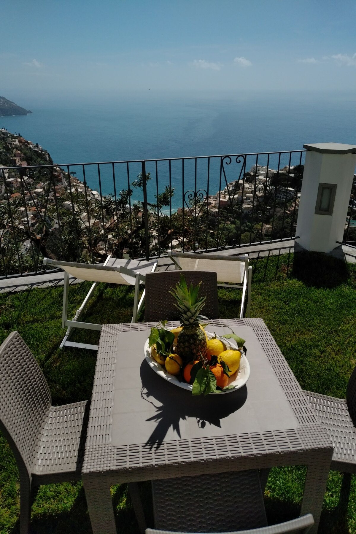 A patio table surrounded by four chairs displays a large bowl of fresh fruit. In the background, a panoramic view of blue ocean water and distant coastal hills is visible, framed by a wrought iron railing. The space is situated on a grassy area, enhancing outdoor dining.