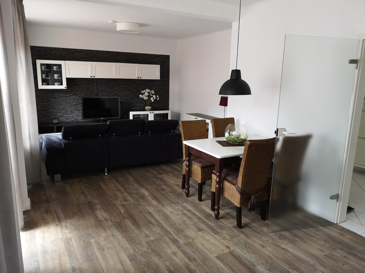 A living area with a dark upholstered sofa faces a wall-mounted television beneath a simple display shelf. A small dining table with wicker chairs is positioned near a glass door, allowing natural light to enhance the space. Light wood flooring complements the neutral color scheme.