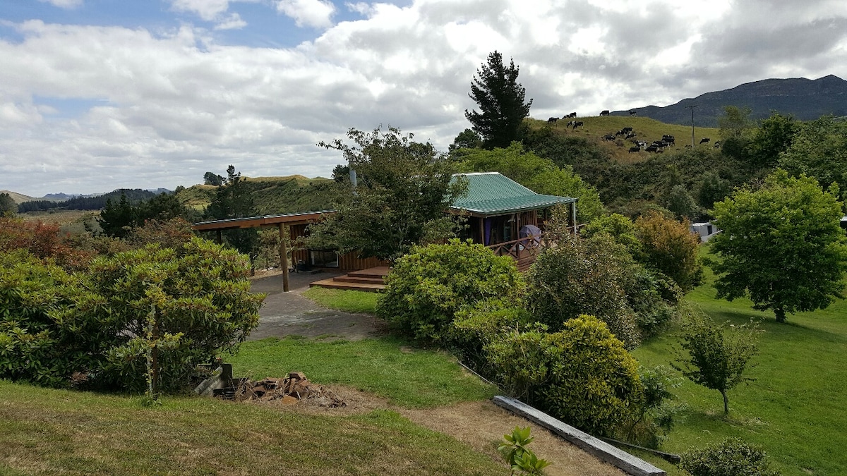 A rustic cottage with a green roof is visible among lush greenery and trees. The outdoor deck extends towards a spacious area, offering a picturesque view of the rolling hills and cattle grazing in the background. The scene reflects a serene country setting.